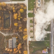Overhead view of industrial building with smoke to the right of houses surrounded by trees with yellow leaves.