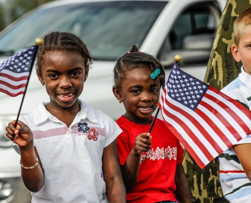 three children in front of white car, two holding an American flag; 4th of July