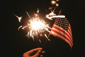 hand holding an American flag and sparkler against dark sky; 4th of July